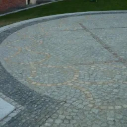 Okrągły plac wyłożony szarą kostką z dekoracyjnym wzorem w kolorze piaskowym, obrzeże z ciemniejszej kostki i jasnego kamienia, fragment muru z czerwonej cegły w tle.