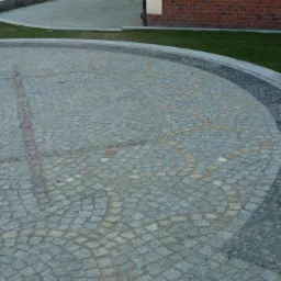 Ułożona kostka granitowa w okrągłym wzorze z elementami dekoracyjnymi w jaśniejszym odcieniu, otoczona obrzeżem z ciemniejszej kostki, widok z góry na fragment posesji.