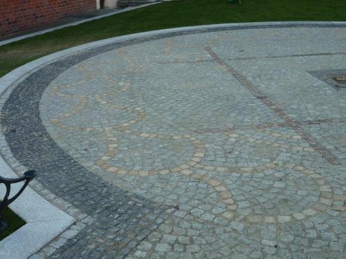 Okrągły plac wyłożony szarą kostką z dekoracyjnym wzorem w kolorze piaskowym, obrzeże z ciemniejszej kostki i jasnego kamienia, fragment muru z czerwonej cegły w tle.