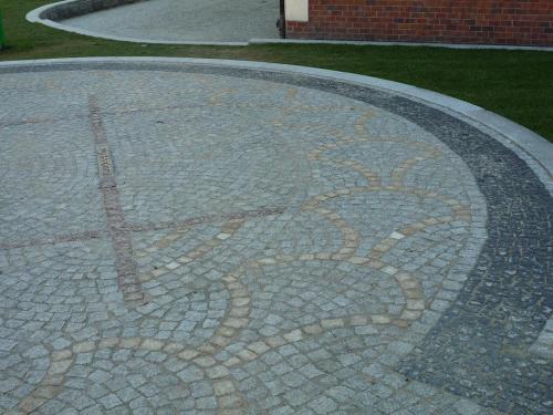 Ułożona kostka granitowa w okrągłym wzorze z elementami dekoracyjnymi w jaśniejszym odcieniu, otoczona obrzeżem z ciemniejszej kostki, widok z góry na fragment posesji.