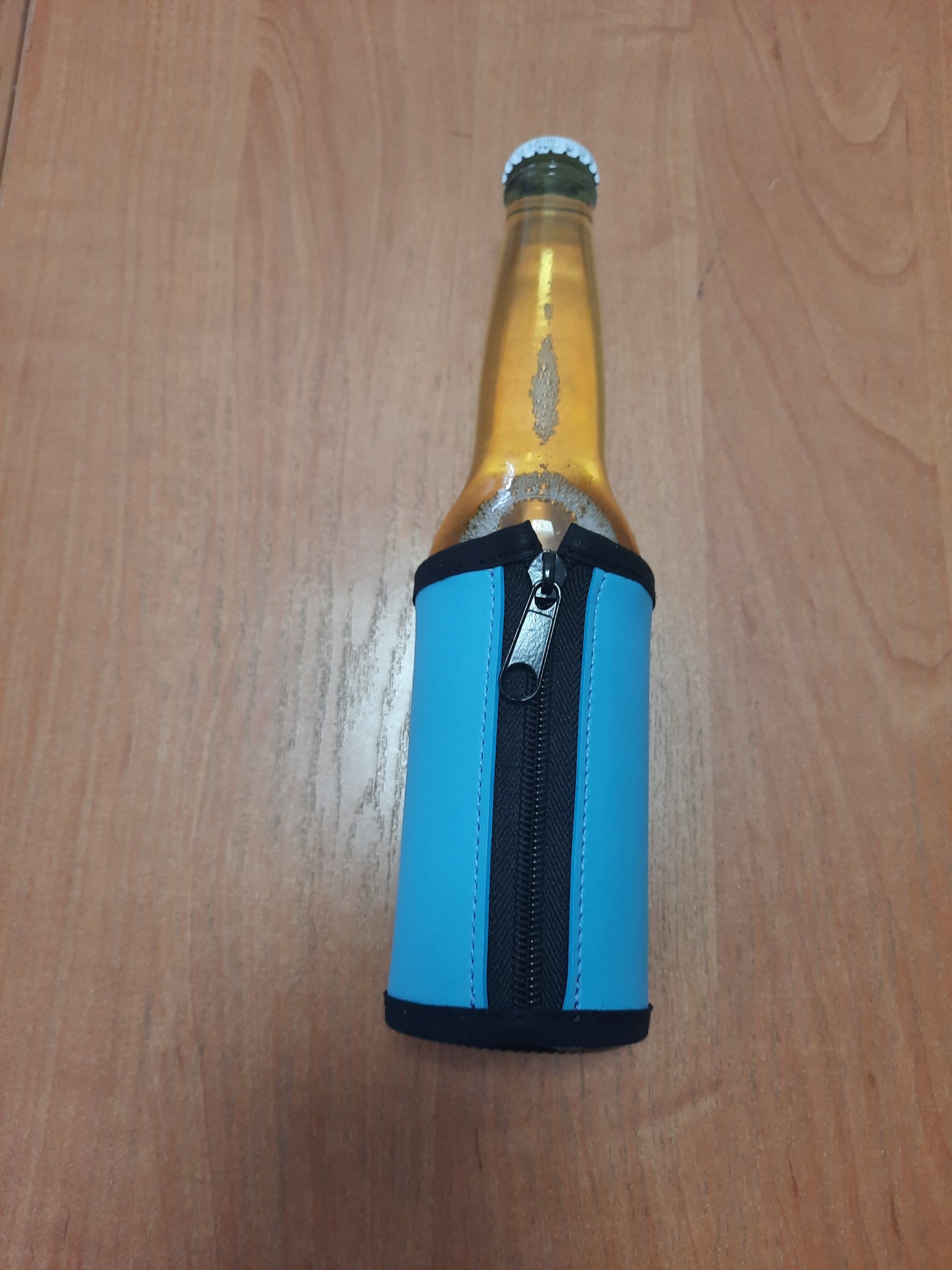 Butelka piwa w turkusowym pokrowcu z zamkiem błyskawicznym, umieszczona na drewnianym stole. Pokrowiec wykonany z dbałością o detale, widoczne przeszycia.