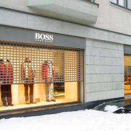ROL-DOM rolety-bramy-żaluzje-markizy - Zewnętrzna, metalowa roleta antywłamaniowa w kolorze szarym, zabezpieczająca witrynę sklepu odzieżowego z logo 'BOSS' w zimowy dzień. Widoczne manekiny wewnątrz.