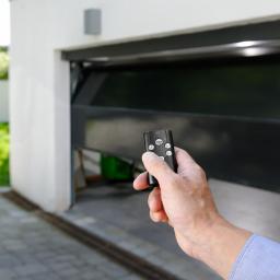 ROL-DOM rolety-bramy-żaluzje-markizy - Otwarta brama garażowa segmentowa sterowana pilotem trzymanym w dłoni, widok z zewnątrz na garaż i fragment ogrodu. Nowoczesne rozwiązanie.