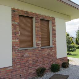 ROL-DOM rolety-bramy-żaluzje-markizy - Dwa okna z brązowymi roletami na ceglanej ścianie budynku jednorodzinnego, widok z zewnątrz, w tle zadbany ogród i kostka brukowa.