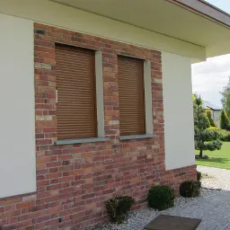 Dwa okna z brązowymi roletami na ceglanej ścianie budynku jednorodzinnego, widok z zewnątrz, w tle zadbany ogród i kostka brukowa.