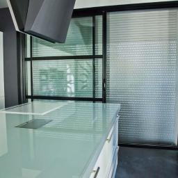 ROL-DOM rolety-bramy-żaluzje-markizy - Nowoczesna kuchnia z przesuwną roletą zewnętrzną w kolorze srebrnym, zapewniającą prywatność i ochronę przed słońcem. Czarna rama okienna, minimalistyczny design.