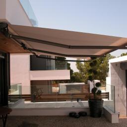 ROL-DOM rolety-bramy-żaluzje-markizy - Beżowa markiza tarasowa z szarym stelażem, zamontowana przy nowoczesnym domu z drewnianym podbitką w Częstochowie. Widok na taras z roślinami.