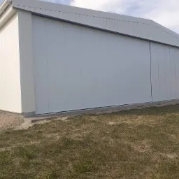 Biały hangar z przesuwną bramą przemysłową, widoczny wiatrowskaz na rogu budynku, trawa na pierwszym planie.