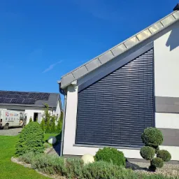 Nowoczesna fasada domu jednorodzinnego z zamontowanymi zewnętrznymi żaluzjami w kolorze grafitowym, widoczny fragment ogrodu i samochód dostawczy firmy montującej z otwartymi drzwiami.