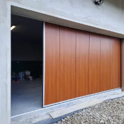 Częściowo otwarta, przesuwna brama garażowa z paneli imitujących drewno, widoczna z zewnątrz na tle surowego betonu i kamiennego podłoża.