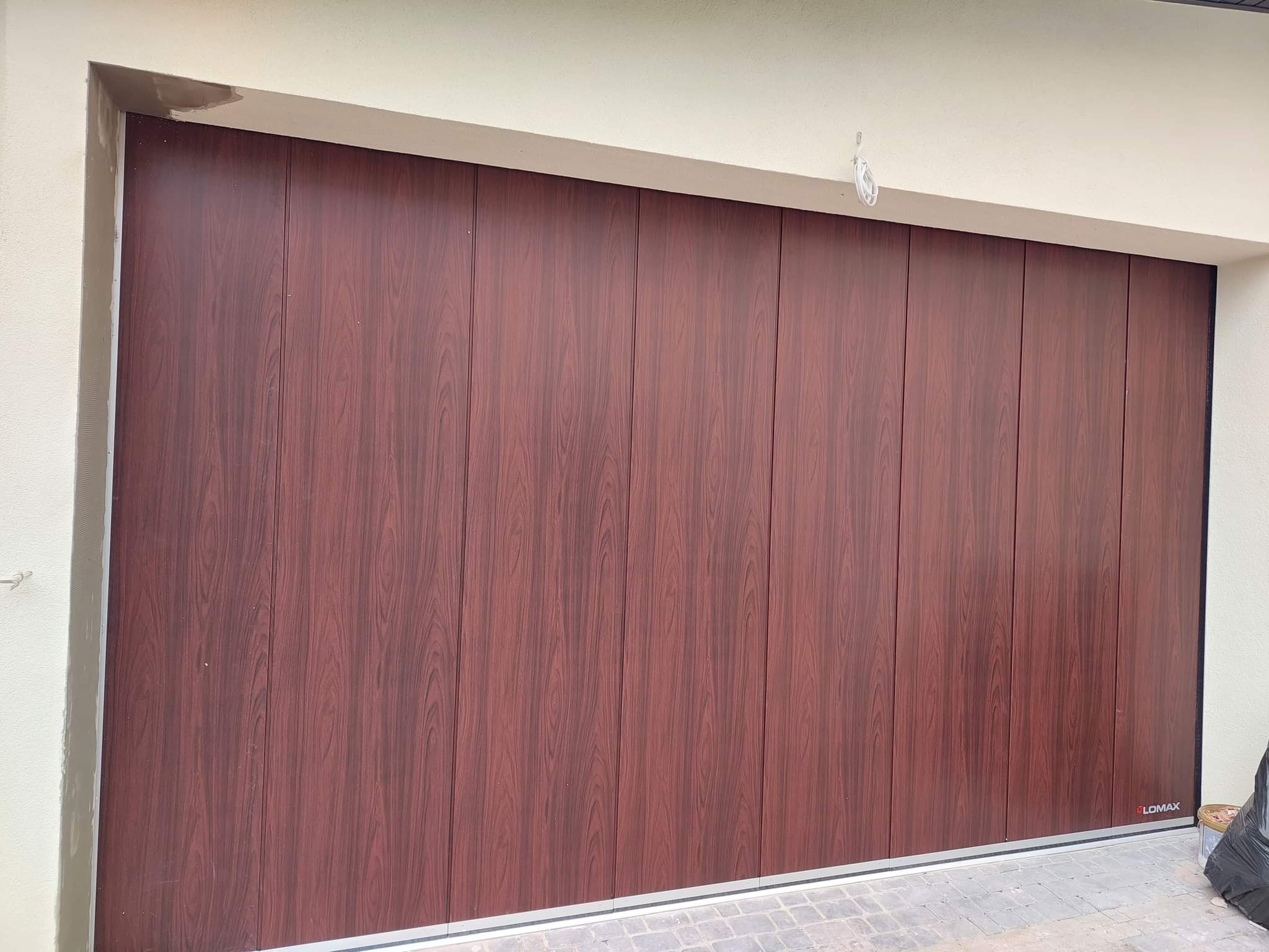 Zamknięta segmentowa brama garażowa z paneli imitujących ciemne drewno, widoczna z zewnątrz, z logo producenta w prawym dolnym rogu.