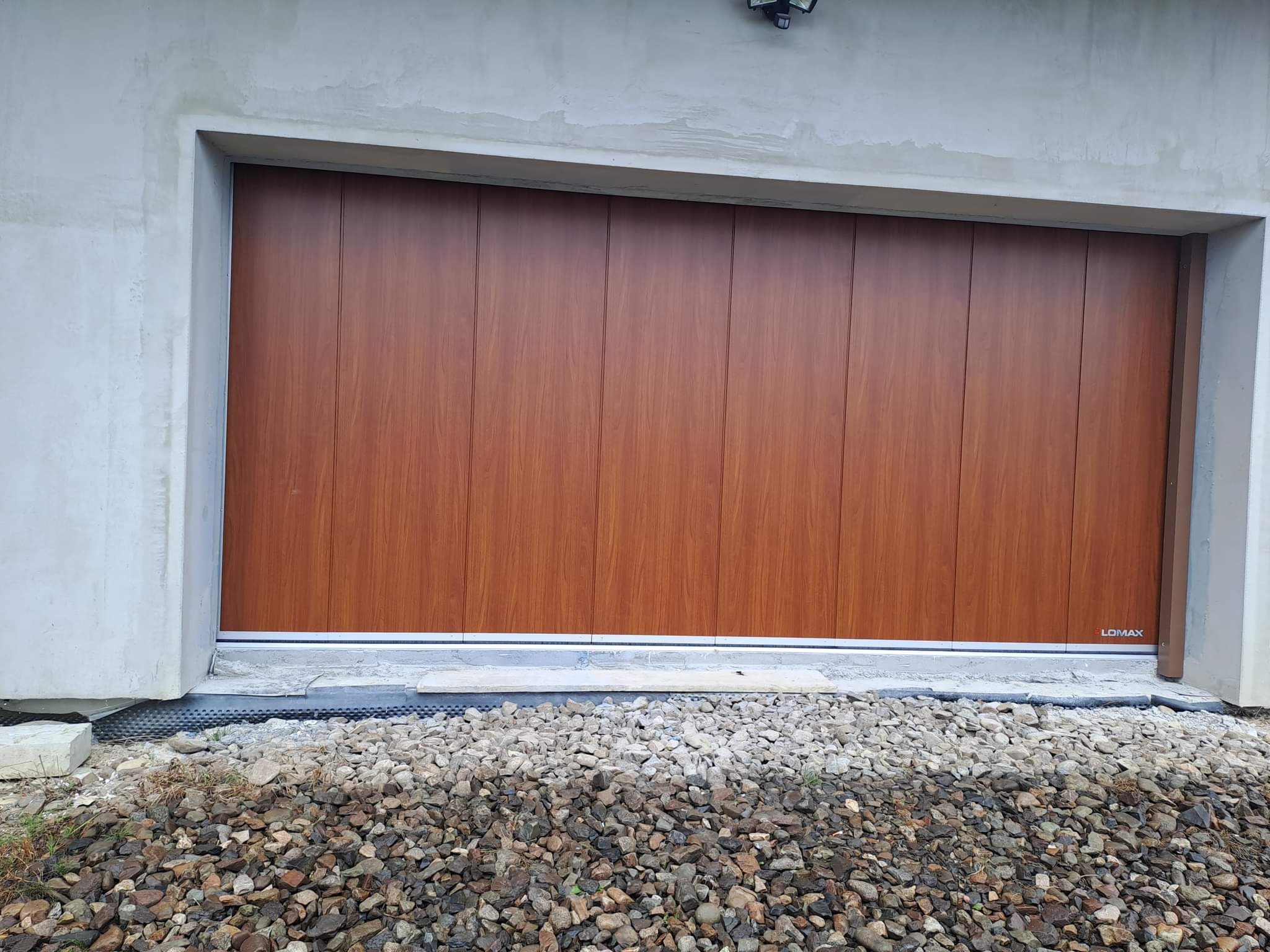 Segmentowa brama garażowa w kolorze drewna, zamontowana w surowym, szarym otworze budynku, z widocznym fundamentem i kamienistym podłożem.