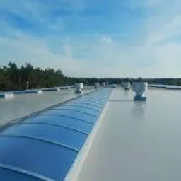 Biała, płaska powierzchnia dachu przemysłowego z niebieskimi świetlikami łukowymi i odpowietrznikami, widok z góry w słoneczny dzień.