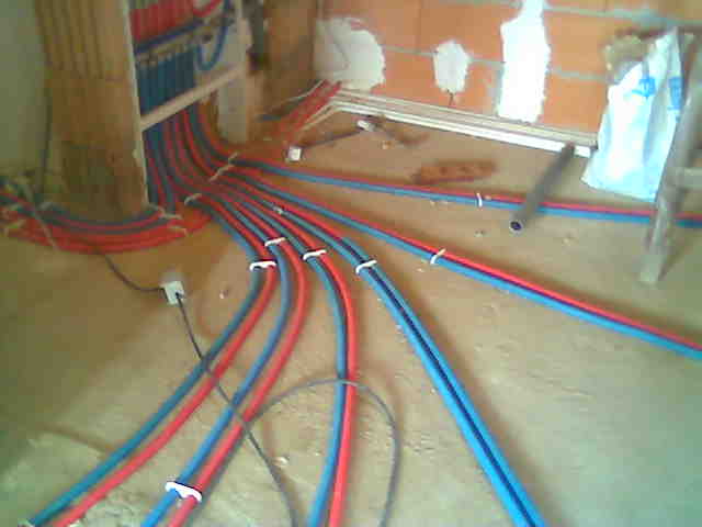 Rozprowadzenie instalacji grzewczej i wodnej w nowo budowanym domu, widoczne czerwone i niebieskie rury przymocowane do podłoża, ściany w trakcie budowy.