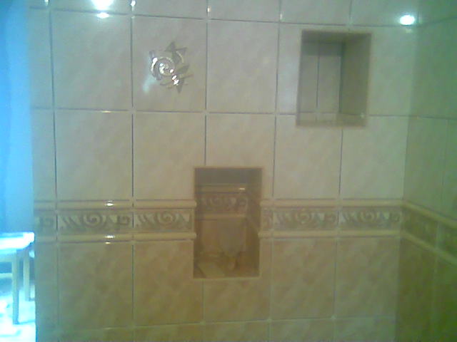 Wykończona ściana łazienki z beżowych płytek ceramicznych z dekoracyjnym motywem roślinnym i wnękami na akcesoria, widoczne fragmenty wyposażenia łazienki.