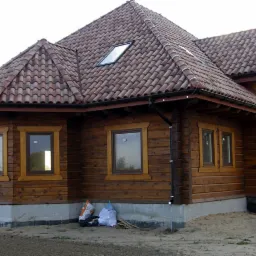 Zewnętrzny widok domu z bali z czerwoną dachówką, widoczne okna z drewnianymi ramami, fundamenty i teren wokół budynku.