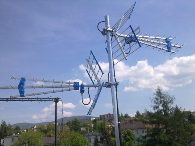 Złożona antena satelitarna z niebieskimi elementami, zamontowana na dachu budynku z widokiem na miasto i błękitne niebo z chmurami.