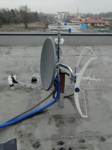 Antena satelitarna i antena radiowa zamontowane na płaskim dachu budynku, z widocznymi przewodami i elementami mocującymi.