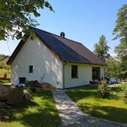 Parterowy dom z białym tynkiem i ciemną dachówką, widok z boku na ogród z kostką brukową, drzewami i stertą drewna.