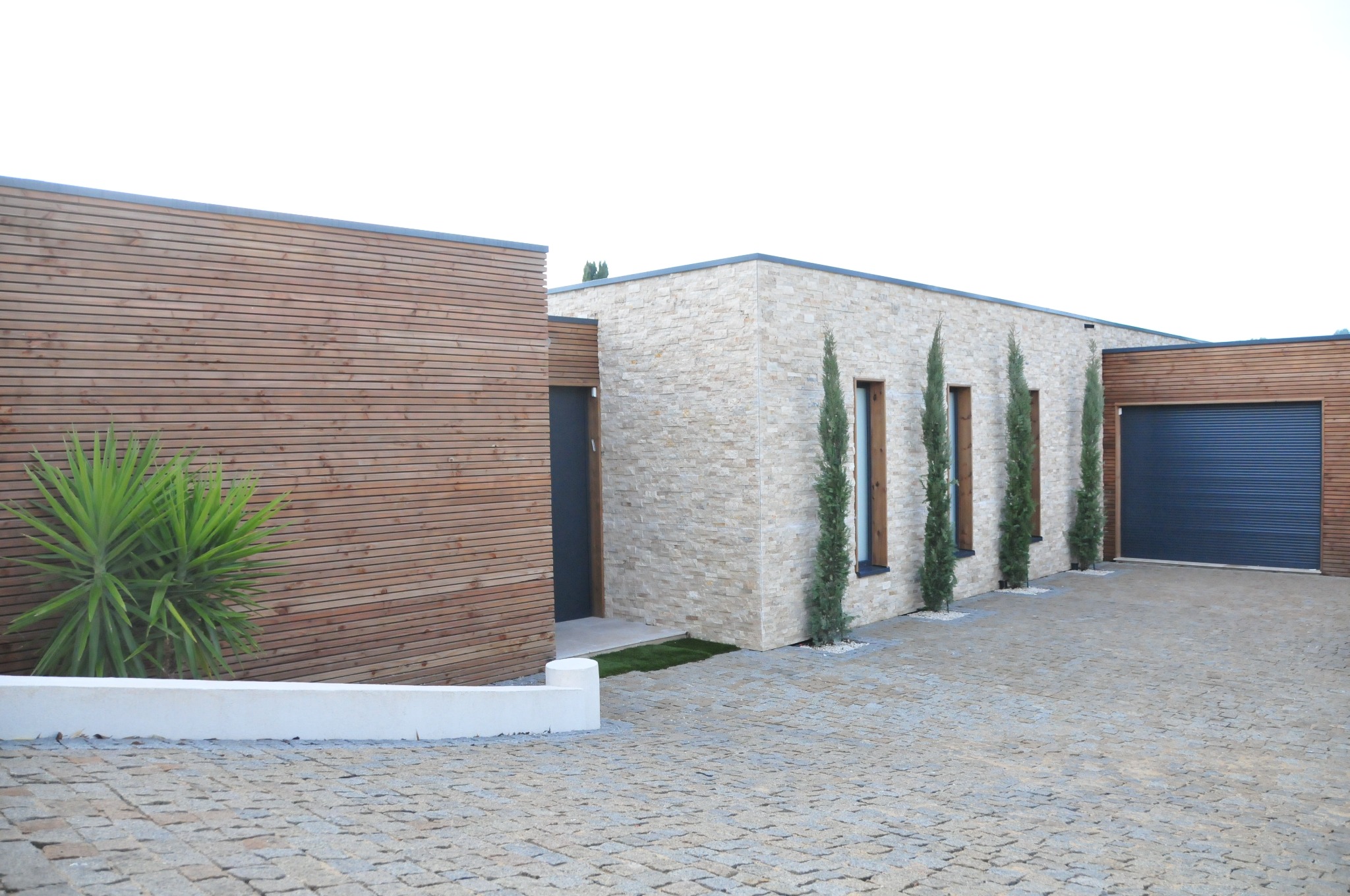 Nowoczesny parterowy dom z elewacją z jasnego kamienia i drewna, trzy smukłe cyprysy przed oknami, podjazd wyłożony kostką brukową, egzotyczna roślina przy drewnianej ścianie.