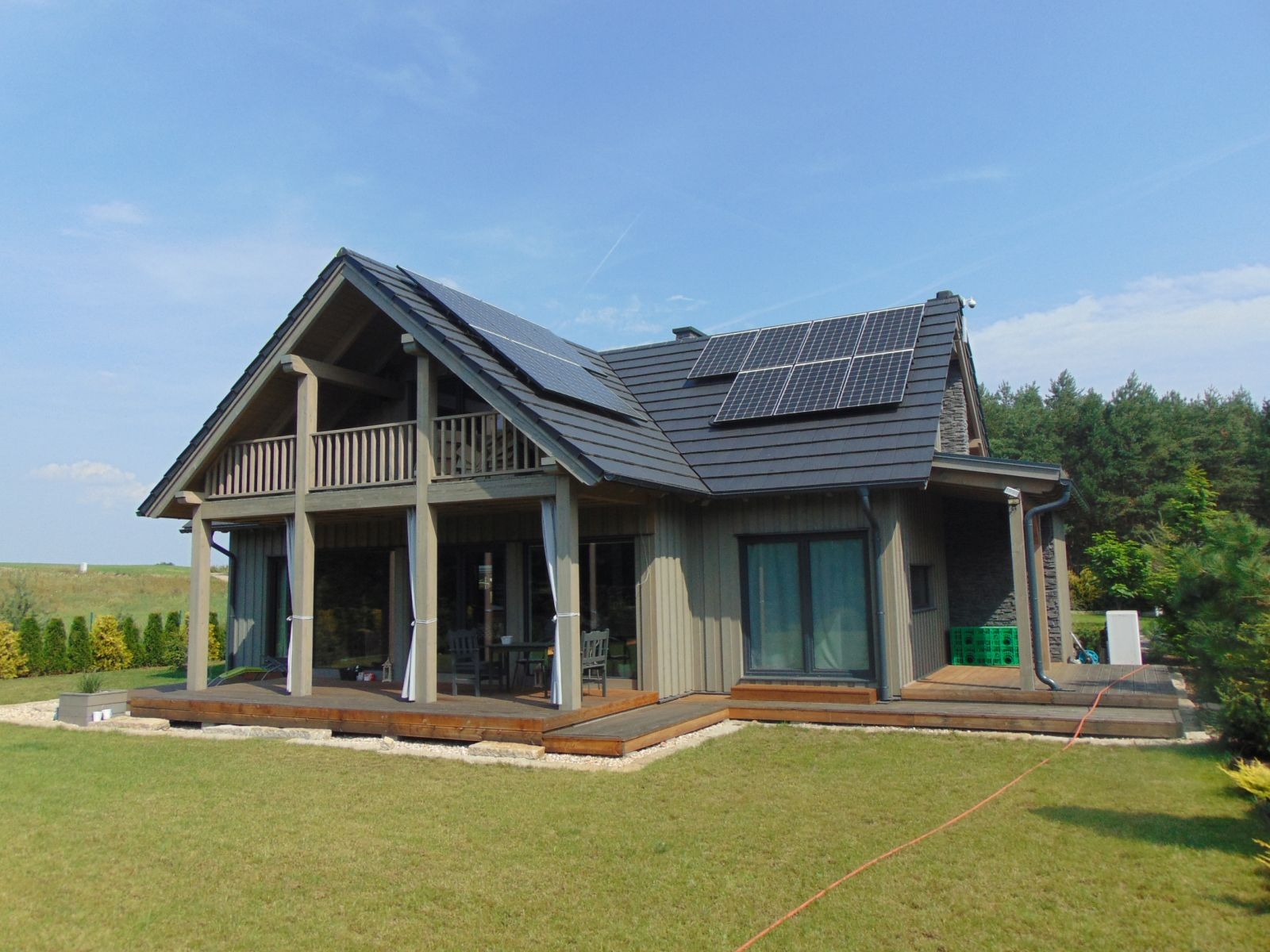 Nowoczesny dom z drewnianą elewacją, balkonem i panelami słonecznymi na dachu, otoczony zielonym trawnikiem i drzewami.