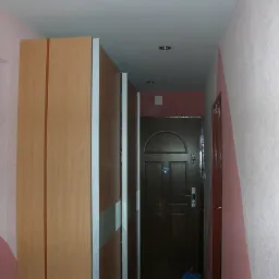 Wnętrze przedpokoju z szafą przesuwną o drewnianym wykończeniu, ciemnymi drzwiami wejściowymi i ścianami pomalowanymi na biało i różowo, oświetlenie punktowe na suficie.