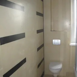 Wykończona łazienka z beżowymi płytkami i poziomymi czarnymi pasami dekoracyjnymi na ścianie, widoczna toaleta podwieszana z chromowanym przyciskiem spłukiwania i fragment kabiny prysznicowej.