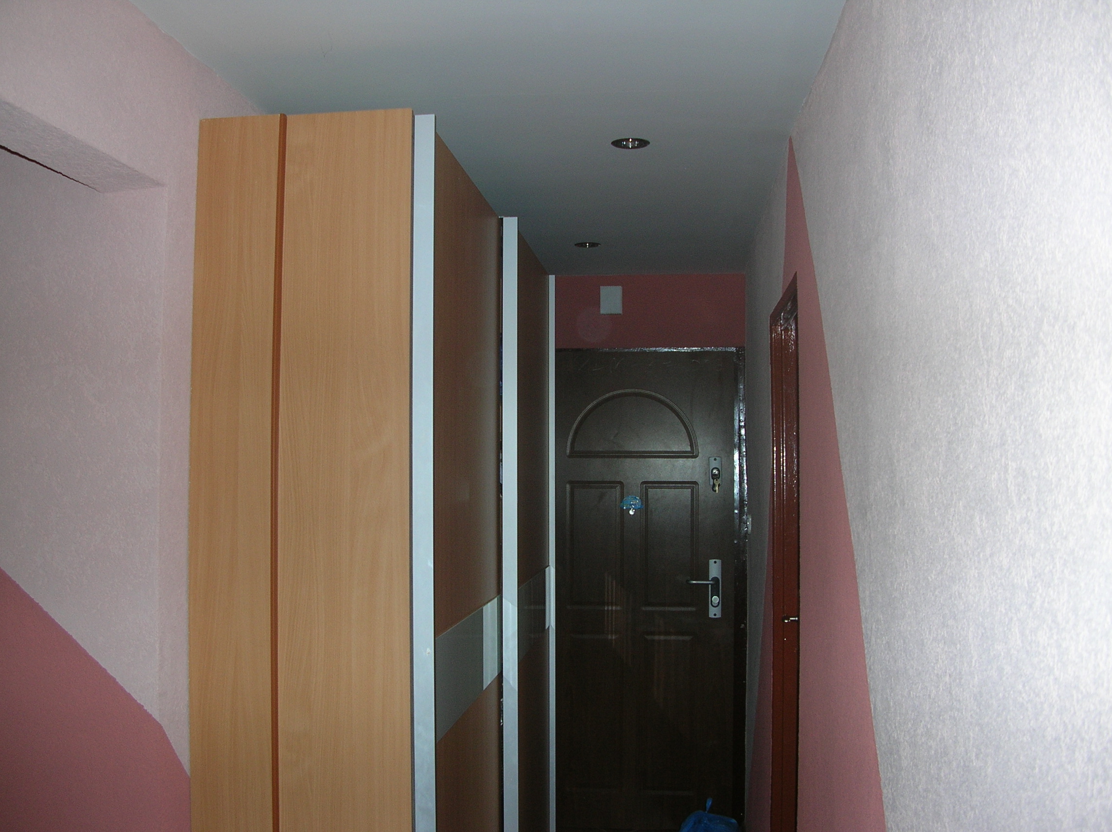 Wnętrze przedpokoju z szafą przesuwną o drewnianym wykończeniu, ciemnymi drzwiami wejściowymi i ścianami pomalowanymi na biało i różowo, oświetlenie punktowe na suficie.
