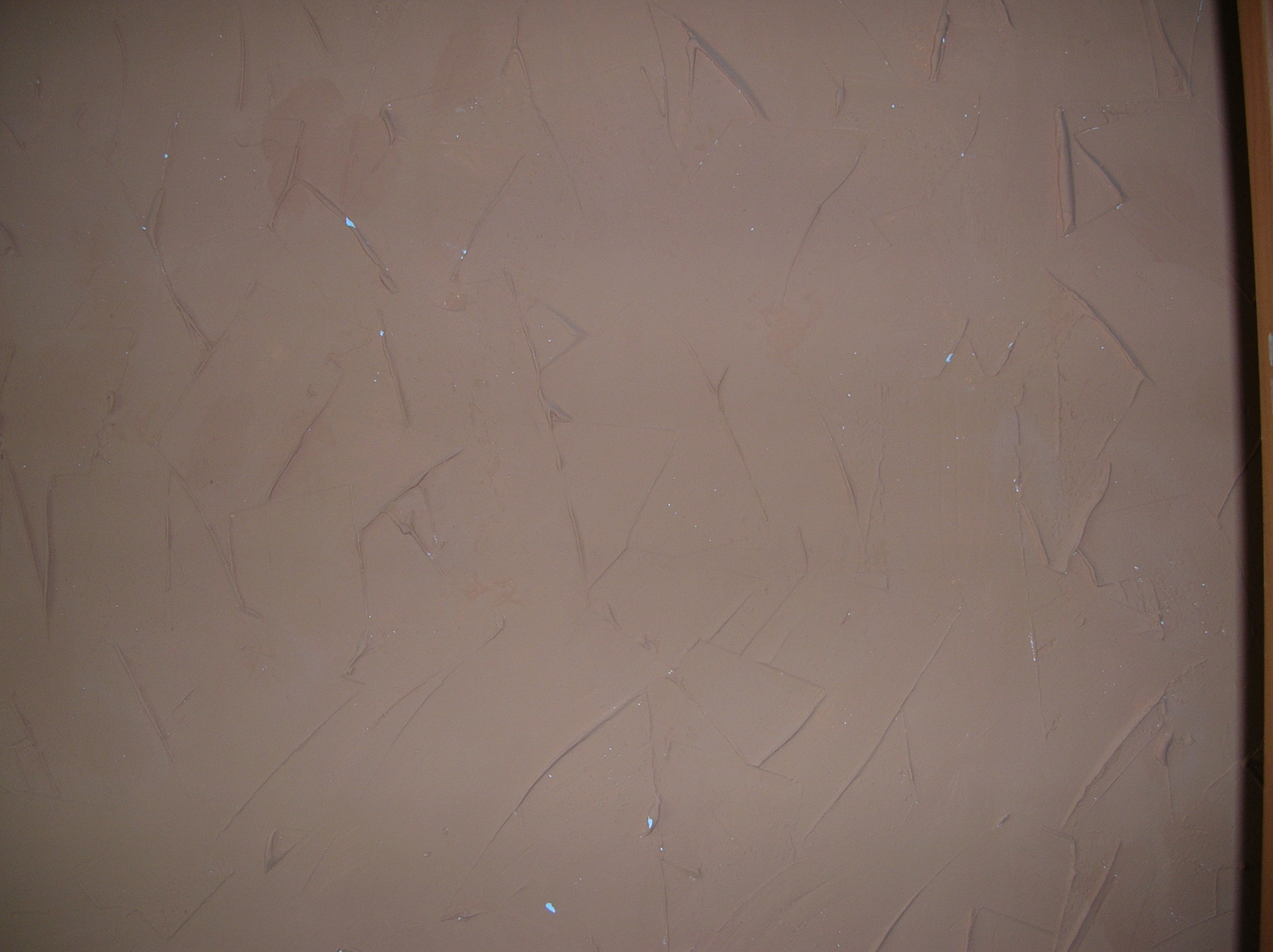 Brązowa ściana z nałożoną fakturą, widoczne ślady narzędzi i drobne niedoskonałości, fragment futryny po prawej stronie.