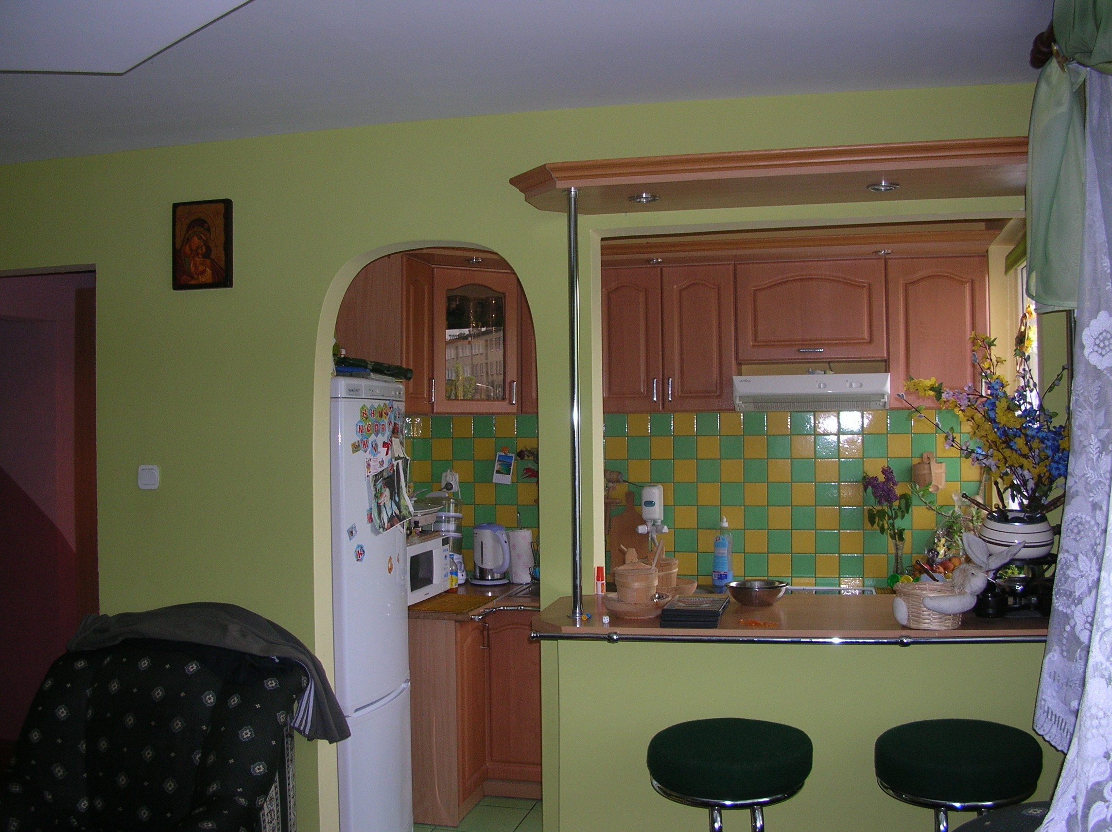 Widok na kuchnię z charakterystycznymi zielono-żółtymi kaflami na ścianie, drewnianymi szafkami, półwyspem z dwoma stołkami i dekoracjami, w tym bukietem kwiatów i obrazkiem religijnym na ścianie.