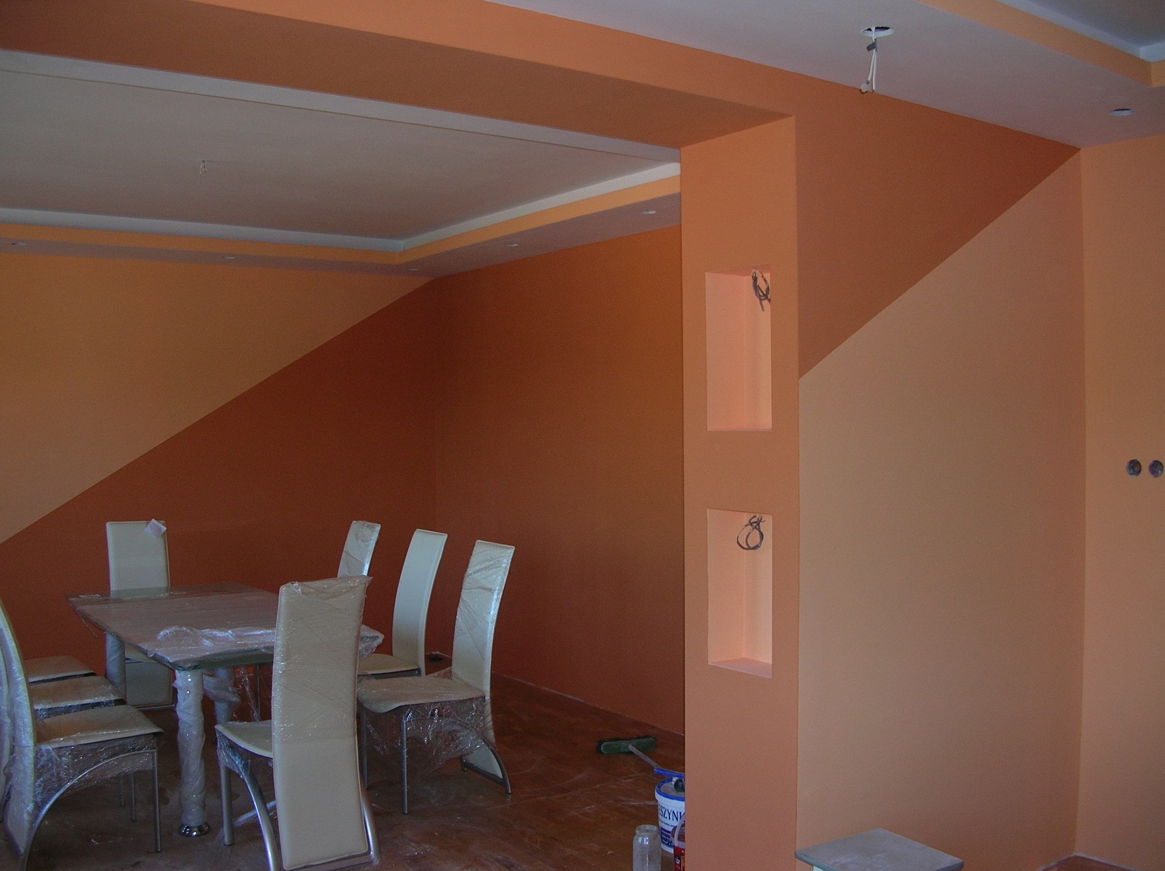 Wnętrze pomalowane na dwa odcienie brzoskwini, z widocznymi niedokończonymi instalacjami elektrycznymi, zabezpieczonymi folią meblami i wnękami w ścianie.