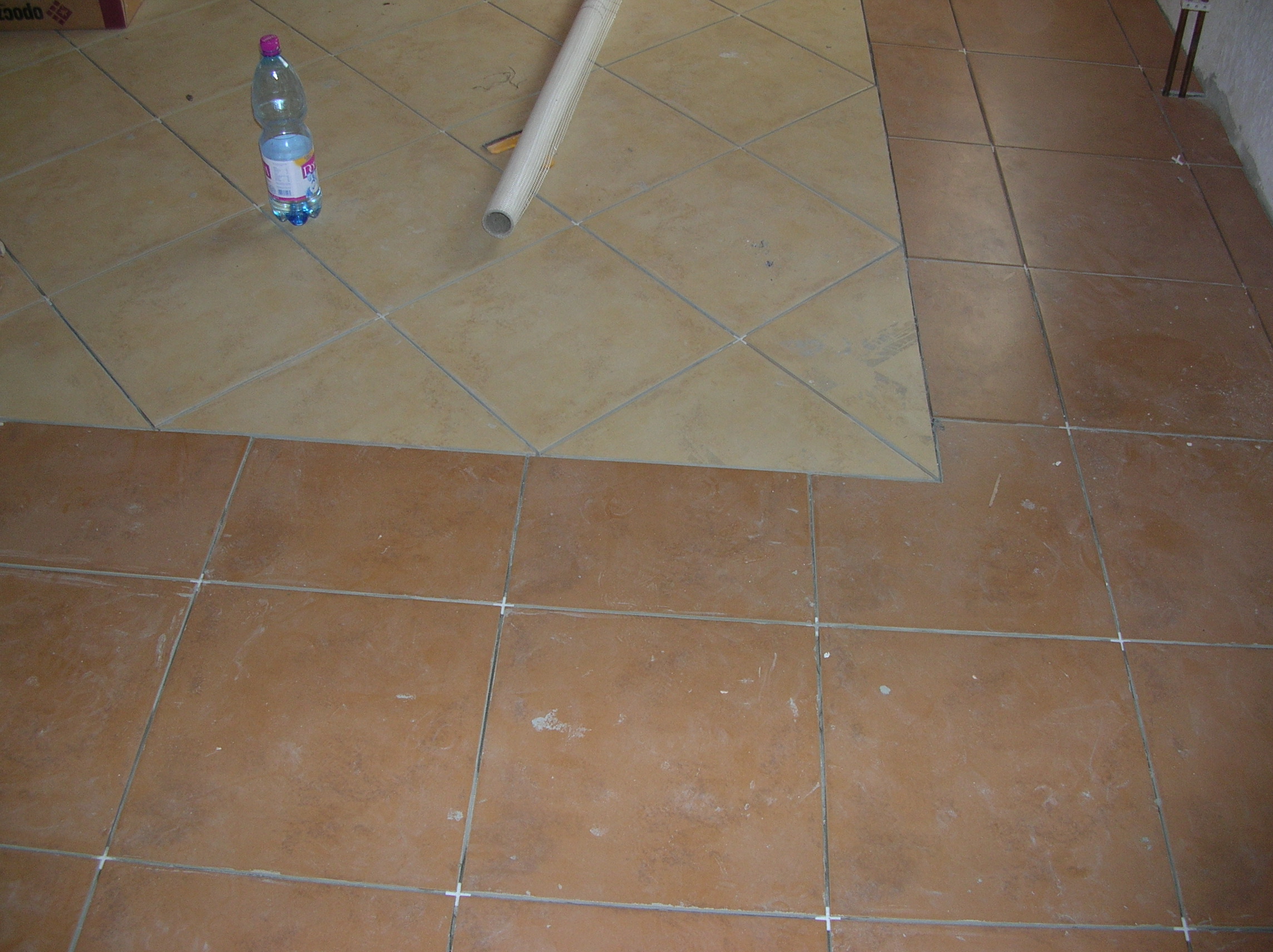 Fragment podłogi z ułożonymi częściowo brązowymi i beżowymi płytkami, widoczna butelka wody i zwinięty materiał budowlany, krzyżyki dystansowe między płytkami.