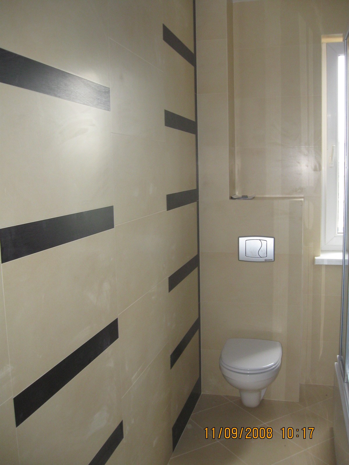 Wykończona łazienka z beżowymi płytkami i poziomymi czarnymi pasami dekoracyjnymi na ścianie, widoczna toaleta podwieszana z chromowanym przyciskiem spłukiwania i fragment kabiny prysznicowej.