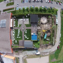 Widok z góry na plac budowy z częścią rekreacyjną: wznoszona konstrukcja szkieletowa obok basenu i terenu z leżakami, z żółtym dźwigiem w pobliżu.