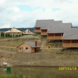 Kompleks drewnianych domków rekreacyjnych z szarymi dachami, usytuowanych na wzgórzu nad stawem, z widocznym domem o nietypowej, okrągłej konstrukcji dachu w tle.