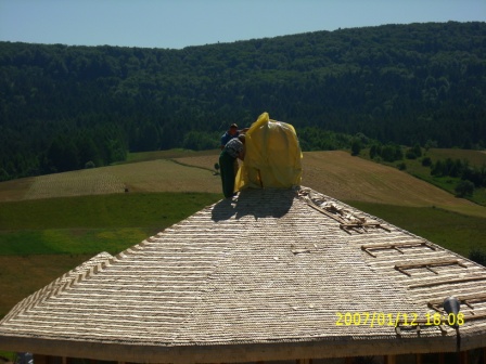 Dwóch mężczyzn pracuje na okrągłym dachu pokrytym strzechą, zabezpieczając komin żółtą folią na tle pagórkowatego krajobrazu z lasami.