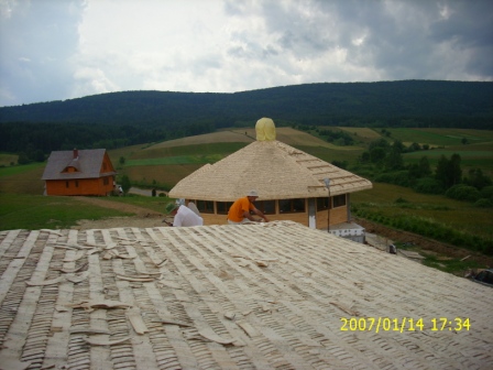 Ręczne krycie strzechą dachu okrągłego budynku z drewnianą konstrukcją, widok z dachu pokrytego papą, w tle krajobraz zalesionych wzgórz i drewniany dom.