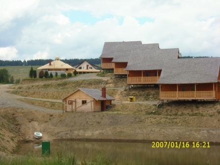 Kompleks drewnianych domków rekreacyjnych z szarymi dachami, usytuowanych na wzgórzu nad stawem, z widocznym domem o nietypowej, okrągłej konstrukcji dachu w tle.