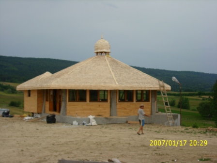Budowa okrągłego, drewnianego domu z nietypowym dachem krytym strzechą, widoczny mężczyzna w krótkich spodenkach, drabina oparta o ścianę budynku, w tle zalesione wzgórza.