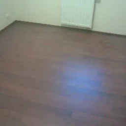 Ułożona, brązowa podłoga panelowa w pustym pokoju z białymi ścianami i grzejnikiem, widok z góry, brak listew przypodłogowych.