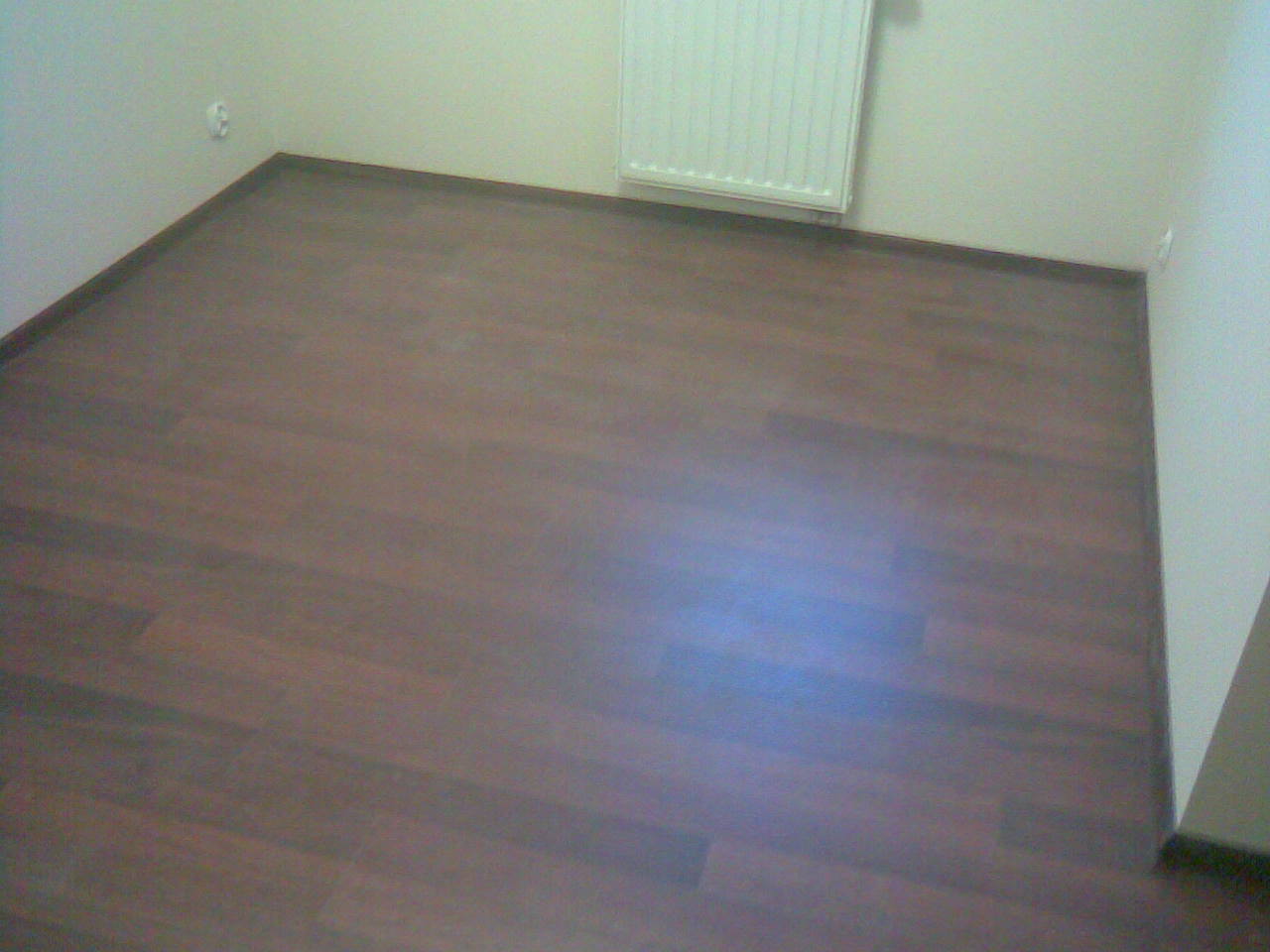 Ułożona, brązowa podłoga panelowa w pustym pokoju z białymi ścianami i grzejnikiem, widok z góry, brak listew przypodłogowych.