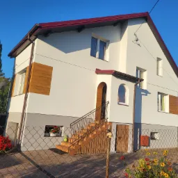 Dwukondygnacyjny dom z białymi ścianami, szarą podmurówką i elementami drewnianej okładziny, widoczny zza metalowej siatki ogrodzeniowej w słoneczny dzień.