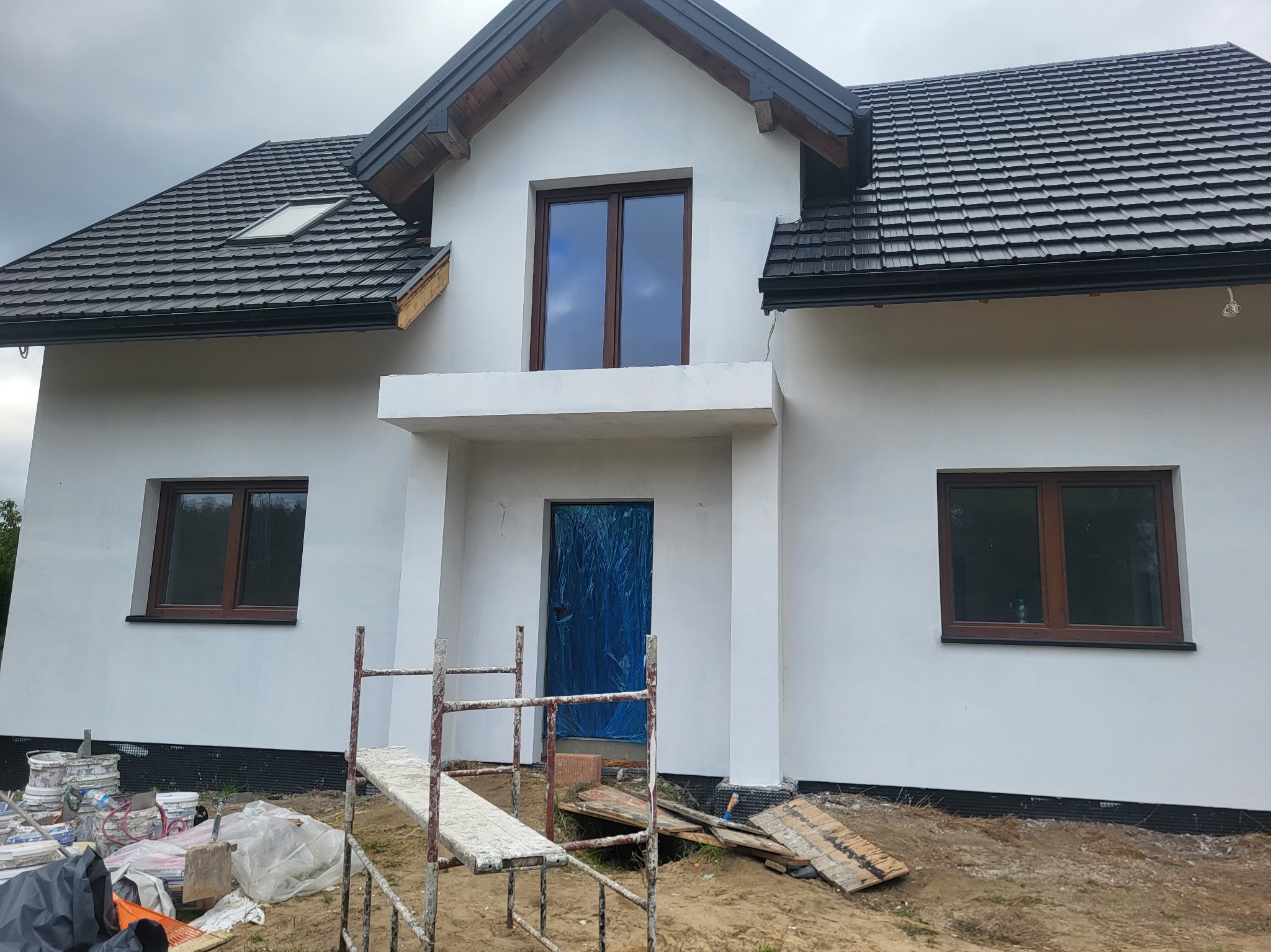 Nowo otynkowany, biały dom jednorodzinny z ciemną dachówką i brązowymi oknami. Drzwi wejściowe zabezpieczone folią. Widoczne rusztowanie i pozostałości materiałów budowlanych.