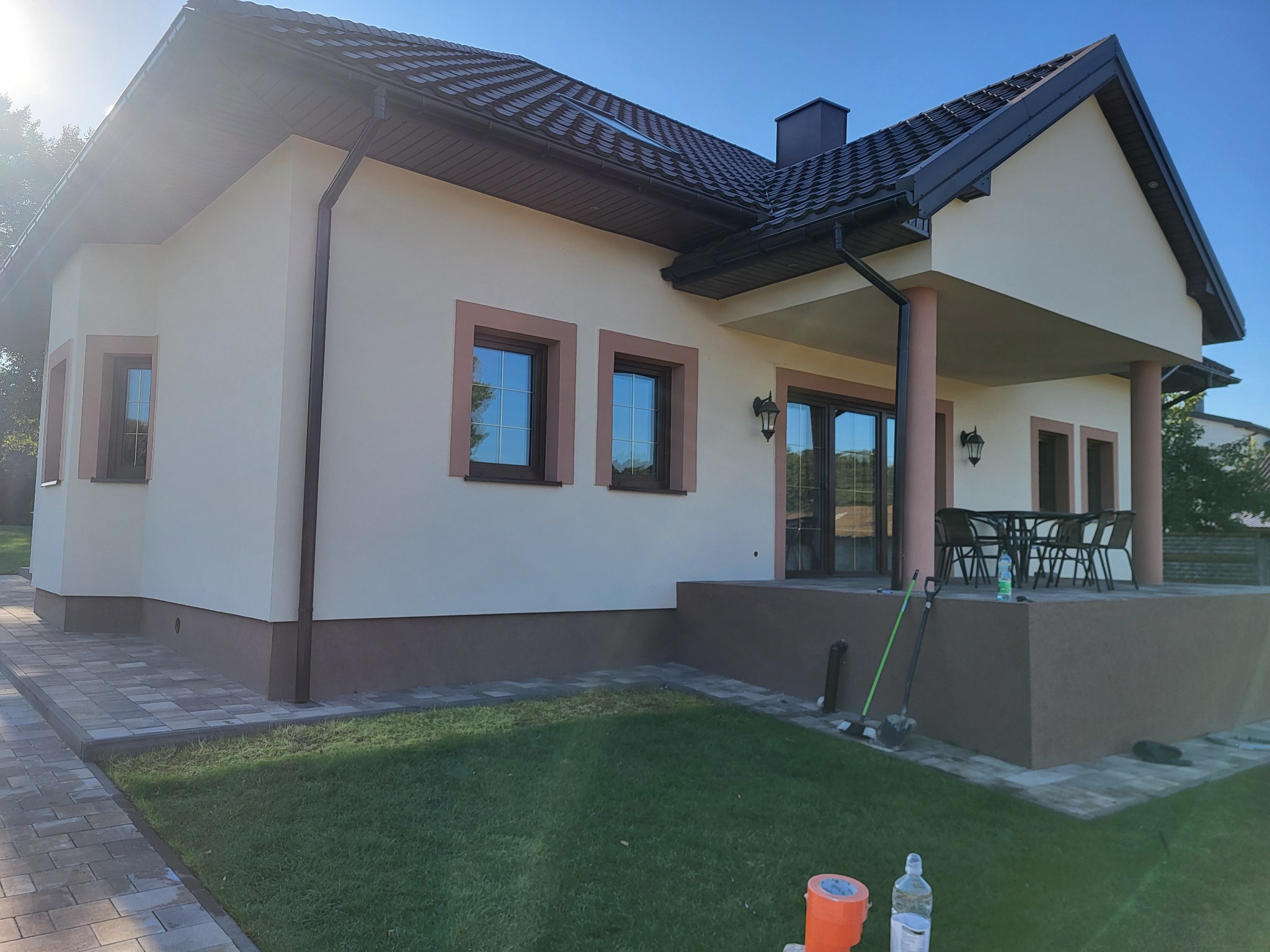 Jasny, jednopiętrowy dom z ciemną dachówką, widoczne detale wykończenia elewacji, okna z brązowymi ramami, taras z meblami ogrodowymi, częściowo zagospodarowany teren wokół domu.