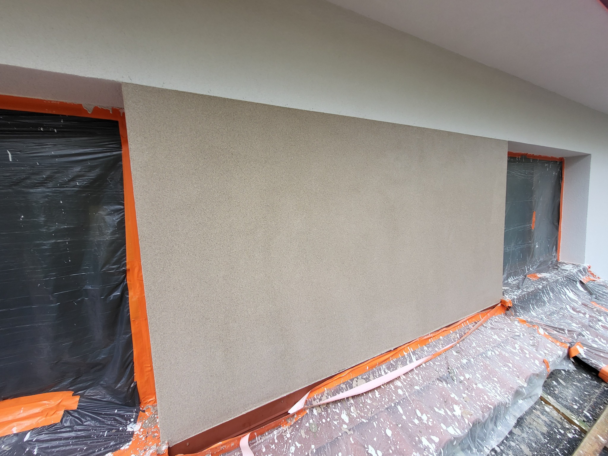 Świeżo nałożony, beżowy tynk strukturalny na ścianie zewnętrznej budynku, zabezpieczone okna folią i taśmą malarską, widoczne ślady zaprawy na folii ochronnej pod ścianą.