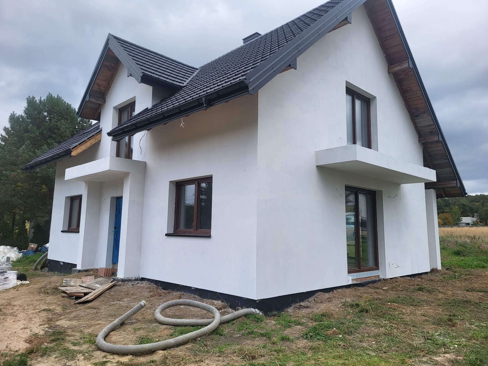 Nowoczesny dom jednorodzinny z białymi ścianami zewnętrznymi i grafitowym dachem, widoczne elementy przygotowania podłoża wokół budynku, wąż do pompy na pierwszym planie.