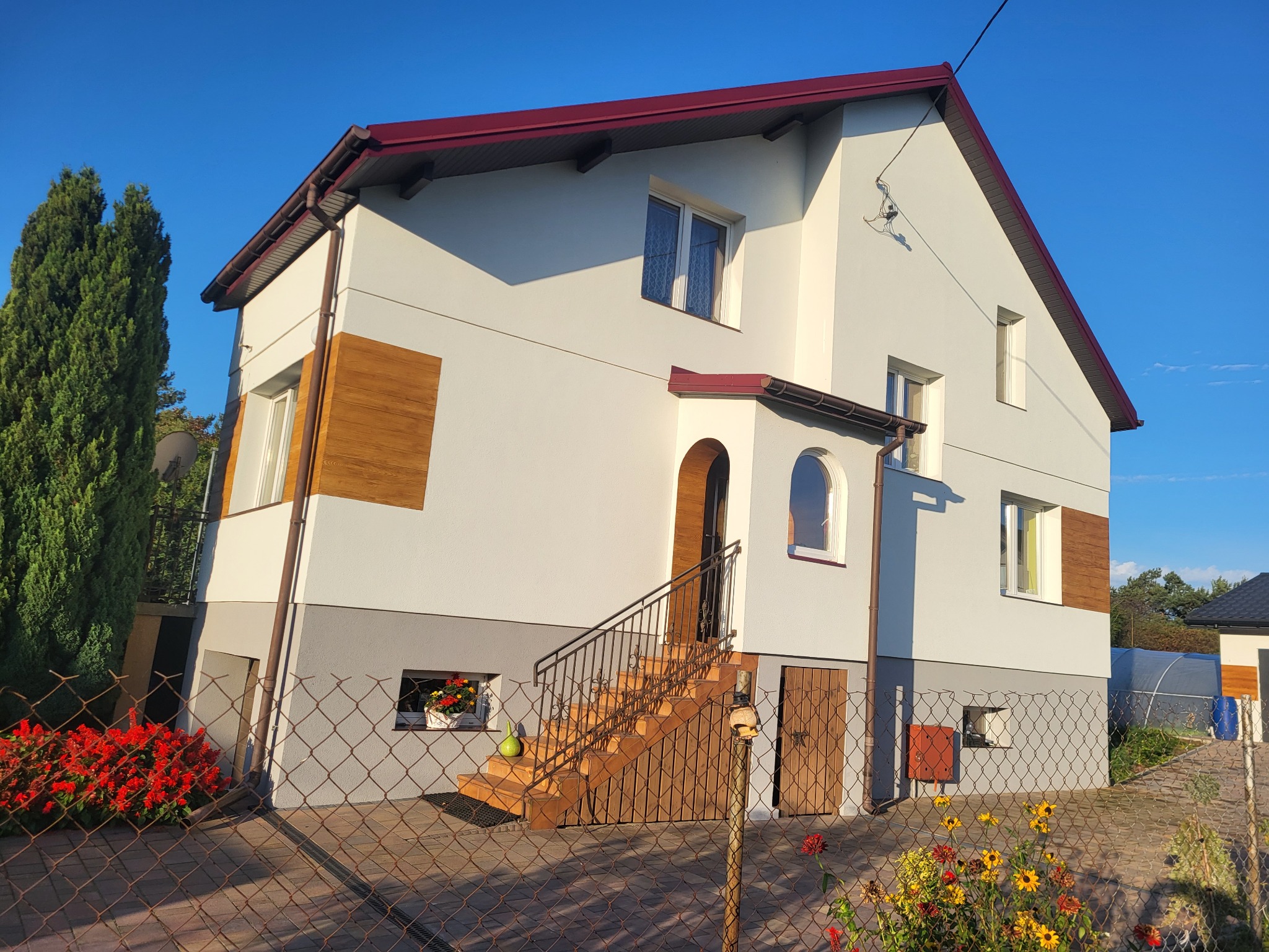 Dwukondygnacyjny dom z białymi ścianami, szarą podmurówką i elementami drewnianej okładziny, widoczny zza metalowej siatki ogrodzeniowej w słoneczny dzień.