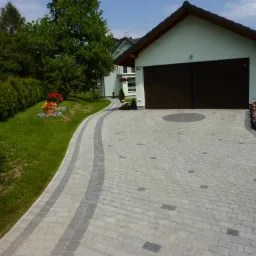 Nowa, jasnoszara kostka brukowa ułożona na podjeździe do garażu, z ciemniejszymi pasami po bokach i okrągłym wzorem przed bramą, w tle budynek mieszkalny z numerem 32 i zadbany trawnik.