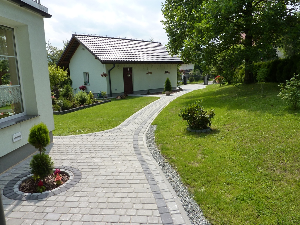 Kaskadowa ścieżka z jasnoszarej kostki brukowej prowadząca do budynku gospodarczego w Biernej, z obrzeżem z ciemniejszej kostki i rabatami z roślinami po bokach, otoczona zielonym trawnikiem.