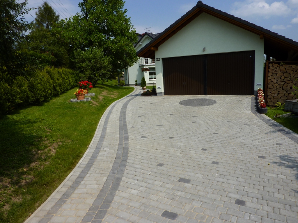Nowa, jasnoszara kostka brukowa ułożona na podjeździe do garażu, z ciemniejszymi pasami po bokach i okrągłym wzorem przed bramą, w tle budynek mieszkalny z numerem 32 i zadbany trawnik.